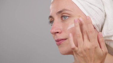 Cilt bakımı reklam konsepti. Stüdyonun gri arka planında kafasında havlu olan güzel bir kadın yüz nemlendirici sürüyor. Sabah güzellik rutini. Makyaj için hazırlık. Boşluğu kopyala