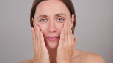 Komik pozitif esmer kadın cilt kremi sürüyor, kameraya özgüvenle bakıyor ve pohpohluyor, yüz gerdirme için nemlendirici krem sürüyor, sağlıklı cilt bakımı için. Stüdyo gri arkaplan