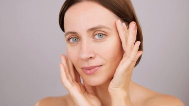 Güzel surat. Yüzüne dokunan mavi gözlü esmer kadın, kameraya bakıyor, stüdyo gri arka planda izole edilmiş. Mükemmel cilde sahip güzel bir kadın model. Cilt bakımı, kozmetik reklamı