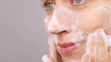 Güzel bir bayanın aşırı yakın çekimi. Temizlik köpüğü süren kadın yüz reklamlarını nazikçe yıkıyor. Baloncuklar. Güzellik rutini. Ticari Temizlik Köpüğü. Cilt bakımı. Uzayı kopyala Gri Arkaplan