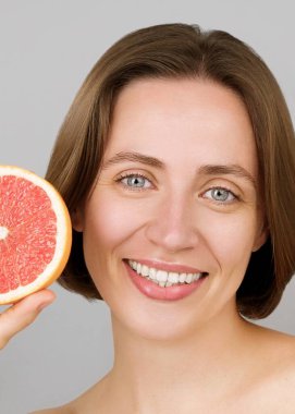 Gülümseyen güzel beyaz model bir greyfurt tutuyor, C vitamini faydaları gösteriyor. Mavi gözlü bir esmerin portresi. Ticari çevre dostu cilt bakımı, detoksifikasyon. Gri arkaplan