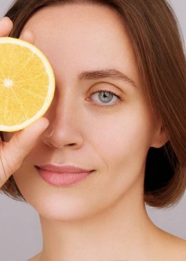 Yakın plan kadın gözlerini portakalı eliyle kesiyor. Sağlıklı ciltli güzel esmer kadın portresi kameraya bakıyor. C vitamini kozmetik konsepti, reklamlar eko cilt bakımı. Gri stüdyo geçmişi