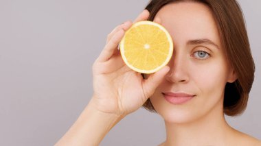 Cilt bakımı. Yakın plan kadın gözlerini portakalı eliyle kesiyor. Güzel esmer kadın portresi, sağlıklı yüz teni kameraya bakıyor. C vitamini kozmetik konsepti, saç bakımı. Gri stüdyo geçmişi
