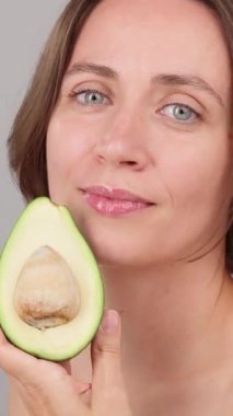 Kafkasyalı sakin bir kadının dikey yakın çekimi yüzünde taze bir avokado tutuyor. Gri arka plan. Ticari cilt bakımı, cilt bakımı, sağlık hizmetleri, organik çevre dostu kozmetik ürünleri ve güzellik merkezi.