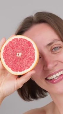 Dikey gülümseyen Kafkasyalı kahverengi model bir gözünü bir greyfurtla kaplıyor ve C vitamini avantajları gösteriyor. Reklam, çevre dostu güzellik, sürdürülebilir cilt bakımı, detoks besin. Gri stüdyo geçmişi