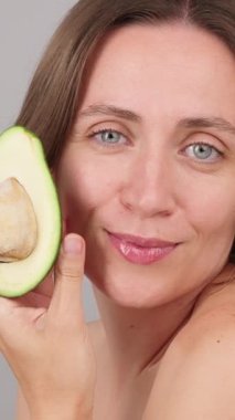 Çekici beyaz bir kadının dikey yavaş çekimde yüzüne taze bir avokado tutarken. Reklamlar, organik çevre dostu kozmetikler, detoks güzellik spa prosedürleri. Gri stüdyo geçmişi