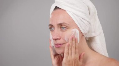 Yüz temizleyici köpük yayılıyor. Yüz yıkama ve gülümseme için nemlendirici köpük kullanan bir kadın, sabah rutini. Stüdyo Gri Arkaplan 'ı izole etti. Güzellik rutini. Cilt bakımı. Boşluğu kopyala.