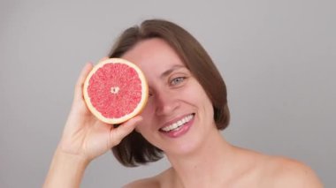 Gülümseyen Kafkasyalı kahverengi model bir gözünü bir greyfurtla kaplayarak C vitamini faydalarını gösteriyor. Çevre dostu güzelliğe, sürdürülebilir cilt bakımına, gri bir stüdyo geçmişine odaklanın.