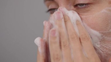 Bir kadın yüzünün aşırı yakın plan çekimi. Temizlik köpüğü süren kadın yüzünü nazikçe yıkıyor. Stüdyo Gri Arkaplan. Günlük güzellik rutini. Reklam köpüğü, cilt bakımı. Kişisel bakım. Boşluğu kopyala