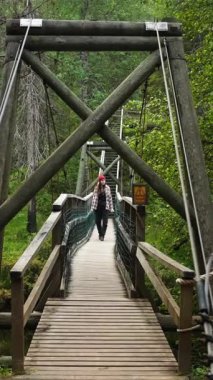 VERTICAL VIDEO Front görüntüsü, genç bir turist kadının geniş açılı görüntüsü, ya da Finlandiya 'da bir orman nehri üzerinde asılı bir köprüde yürüyen kadın yürüyüşçü. Seyahat, keşif, özgürlük, doğa ile bağlantı kavramı