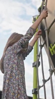 VERTICAL VIDEO gülümseyen okul çağındaki bir kızın modern bir oyun parkında ip merdivene tırmanmasının düşük açılı görüntüsü. Başarı kavramı, eğlence, özgürlük, çocukluk güveni. Açık hava oyunu, aktif yaşam tarzı