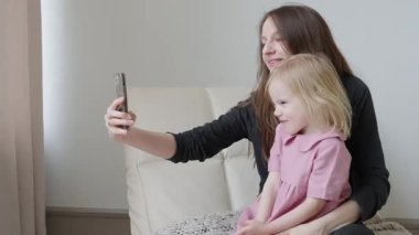 Mutlu anne ve samimi küçük kız evdeki kanepede otururken akıllı telefon, bağlanmak için internet uygulamaları, iş gezisinde babayla görüntülü konuşma. Birlikte selfie çekiyoruz. Video mesajı gönderiliyor.