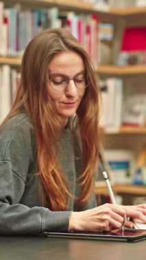 Mutlu ve odaklanmış bir kız öğrenci online bir ders veya ders, araştırma projesi, webinar, sınav hazırlığı için bir üniversite kütüphanesinde notlar alarak dizüstü bilgisayar ve tablet kullanır..