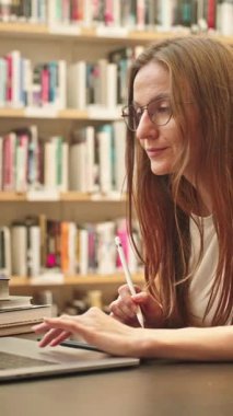 VERTICAL VIDEO: Üniversite kütüphanesinde dizüstü bilgisayar ve internet kullanan genç kız öğrenci, kitaplıklarla çevrili bir masada oturuyor, akademik araştırma yapıyor, notlar alıyor, çevrimiçi öğrenim görüyor.