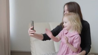 Mutlu anne ve tatlı küçük kızı evdeki koltukta otururken akıllı telefon, bağlanmak için internet uygulamaları, iş gezisinde babayla görüntülü konuşma. Birlikte selfie çekiyoruz. Video mesajı gönderiliyor.