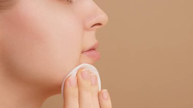 Stüdyoda bej arka planda pamuk ped kullanan gülümseyen genç bir kadının yarı yüz portresi. Beyaz bir kız pamuk kullanarak cildini temizliyor. Cilt bakımı reklamcılığı kavramı. Boşluğu kopyala