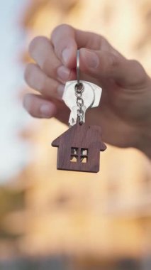 Evin anahtarını elinde tutan genç adamın dikey yakın çekimi ve apartmanın önünde ev şeklindeki anahtarlık. İpotek onayı, konut kredisi, gayrimenkul alımı ve mali bağımsızlık sembolü.