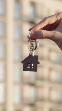 Modern apartman binasının önünde ev şeklindeki anahtarlıkla ev anahtarı tutan kadın emlakçının dikey yakın çekim videosu. Emlak konsepti, mülk sahipliği, ipotek onayı.