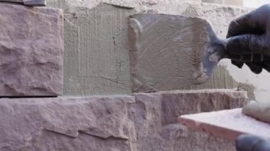 İnşaat işçisi olarak duvar işçiliğinin yakın çekimi, dekoratif taş monte etmeden önce duvara nasıl havan topu atılacağını gösteriyor. Eldivenli eller ön cephe yenileme ve eğitim sırasında yüzeyi hazırlıyor.