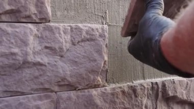 İnşaat işçisi olarak duvar işçiliğinin yakın çekimi, dekoratif taşı taze havan mermisiyle duvara bastırıyor. Ön cephe yenileme ve dış onarım sırasında kaplama yerleştirme koruyucu eldivenler eller.