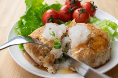 Çeri domatesli ve yeşil yapraklı bir tabağa çatal ve bıçakla kesilmiş tofu Japon hamburgeri bifteği ve üstüne de jöleli turp ve soya sosu.