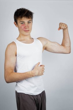 A 17 year old teenage boy wearing a white tank top flexing his arm muscles