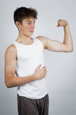 A 17 year old teenage boy wearing a white tank top flexing his arm muscles