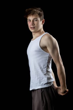 A 17 year old teenage boy wearing a white tank top flexing his arm muscles against a black background