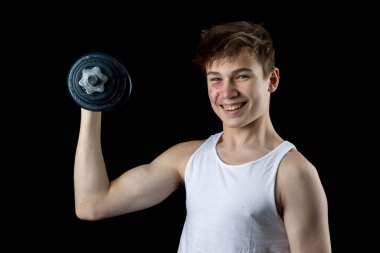 A 17 year old teenage boy wearing a white tank top excercising with a dumbbell against a black background