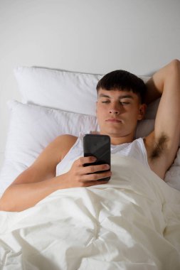 Genç bir erkek, yatakta cep telefonunu kullanıyor.