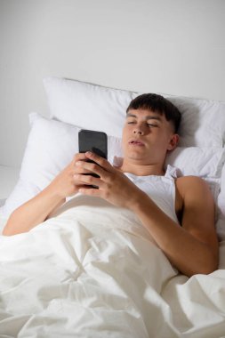 Genç bir erkek, yatakta cep telefonunu kullanıyor.
