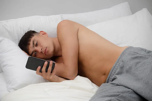 Young single male adult laying shirtless on bed using his phone