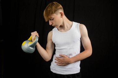 14 yaşında kolsuz yelek giyen beyaz bir çocuğun portresi. Siyah bir arka plan çocuğuna kettlebell kaldırıyor.
