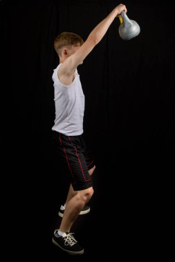 14 yaşında kolsuz yelek giyen beyaz bir çocuğun portresi. Siyah bir arka plan çocuğuna karşı kettlebell salıncakları yapıyor.