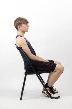 15 yaşında kolsuz tişörtlü ve şortlu bir genç yan yana bir sandalyede oturuyor.