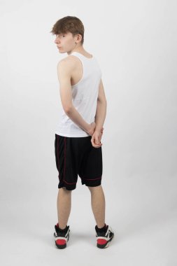 15 yaşında, kolsuz yelek ve şortla beyaz bir arka plana karşı duran ve el ele tutuşup omzuna bakan genç bir çocuk.