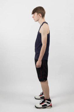 15 yaşında genç bir çocuk kaslı yelek ve şortla beyaz bir arka plana karşı duruyor.