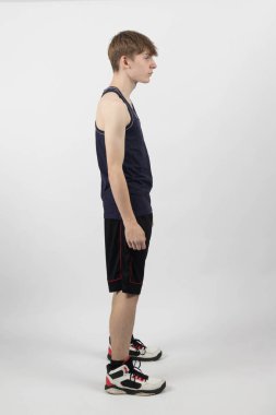 15 yaşında genç bir çocuk kaslı yelek ve şortla beyaz bir arka plana karşı duruyor.