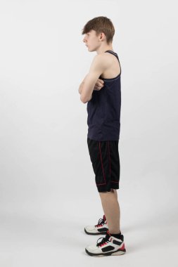 Kas yelekli ve şortlu beyaz bir arka plana karşı duran 15 yaşındaki genç bir çocuk kollarını kavuşturup yan tarafa bakıyor.