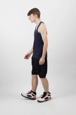 Kas yelekli ve şortlu beyaz bir arka plana karşı duran 15 yaşında bir genç.