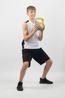 15 yaşında, elinde bir kettlebell olan genç bir çocuk. Stüdyoda beyaz bir arkaplana karşı çekim yapıyor.