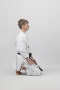 15 yaşında, erkek, karate siyah kuşağı var. Diz çöküyor. Stüdyo çekimi beyaz bir arkaplanda yapılıyor.