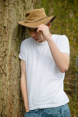 Çalı şapkalı, beyaz tişörtlü ve mavi kotlu genç bir çocuk. Bir el şapkanın üstünde.