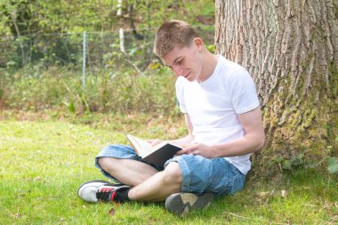 Genç bir çocuk kitap okurken bir ağacın karşısında oturuyor.