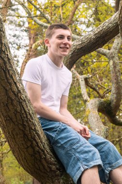 Halka açık bir parkta ağaçta oturan genç bir çocuk, beyaz tişört ve mavi kot pantolon giyiyor.