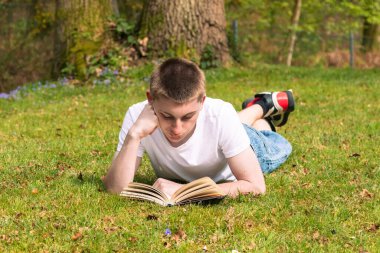 Genç bir çocuk halka açık bir parkta güneşin altında kitap okuyor. Beyaz tişört ve mavi kot pantolon giyiyor.