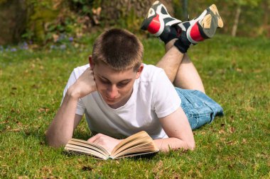 Genç bir çocuk halka açık bir parkta güneşin altında kitap okuyor. Beyaz tişört ve mavi kot pantolon giyiyor.