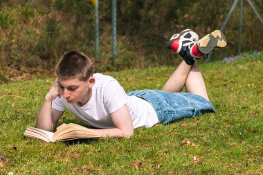 Genç bir çocuk halka açık bir parkta güneşin altında kitap okuyor. Beyaz tişört ve mavi kot pantolon giyiyor.