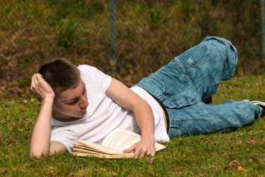 Genç bir çocuk halka açık bir parkta güneşin altında kitap okuyor. Beyaz tişört ve mavi kot pantolon giyiyor.