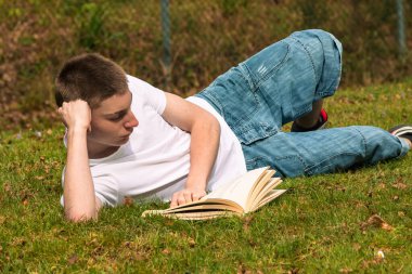 Genç bir çocuk halka açık bir parkta güneşin altında kitap okuyor. Beyaz tişört ve mavi kot pantolon giyiyor.